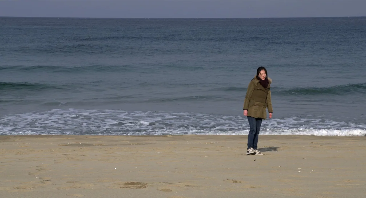 On The Beach At Night Alone (2017) movie still: fullbody — A wide sandy beach stretches toward the calm blue ocean under a clear sky. A young woman…