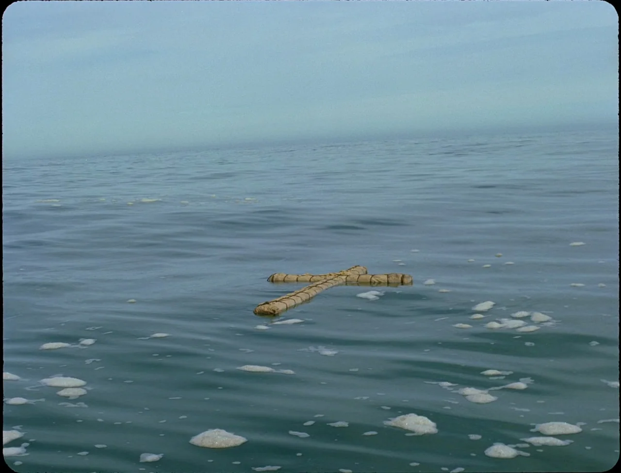Godland (2022) movie still: wide — A cross constructed from bundles of reed or rope drifts on the ocean surface. Small patches of white sea foam…