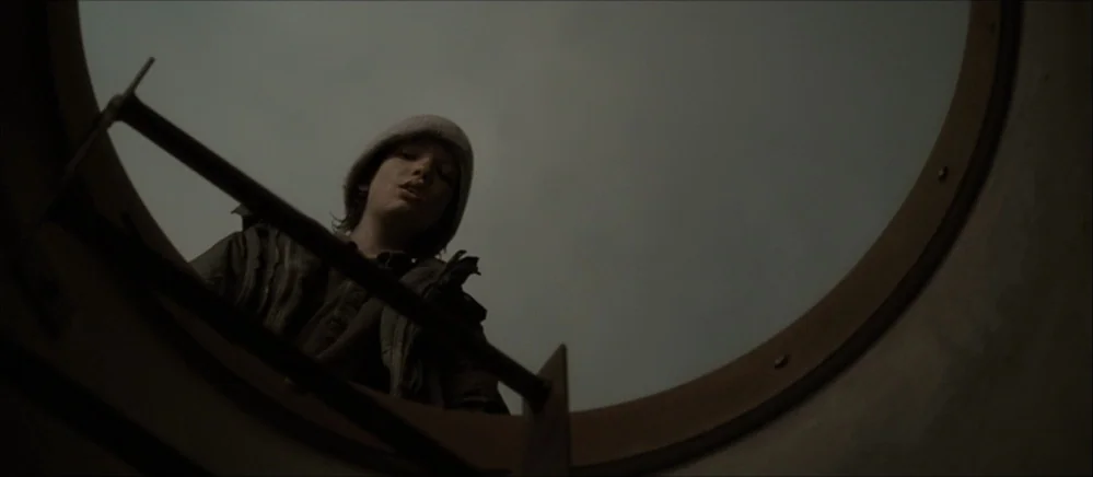 The Road (2009) movie still: medium — A person in a beanie looks down through an open circular steel hatch. The overhead view frames the subject…