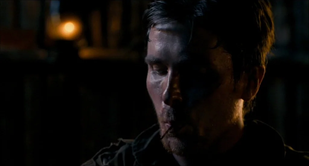 Rescue Dawn movie still: closeup — A dark, dimly lit space with a blurred background showing faint outlines of objects. A man in a dark jacket sits…