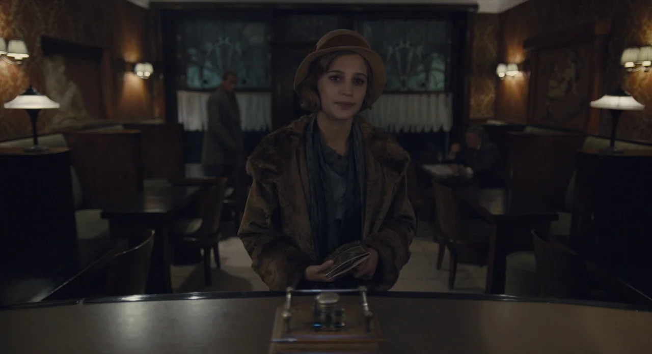 The Danish Girl (2015) movie still: medium — A dimly lit 1920s restaurant interior with dark wooden furniture and patterned wallpaper. A young woman…