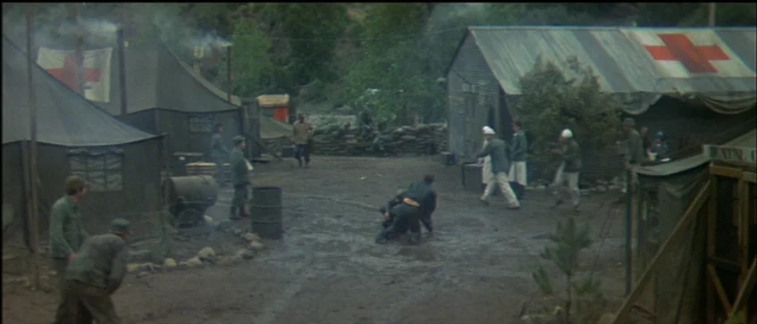 MASH (1970) movie still: wide — A military base camp with tents displaying red cross symbols sits in a clearing. Soldiers in olive drab uniforms…