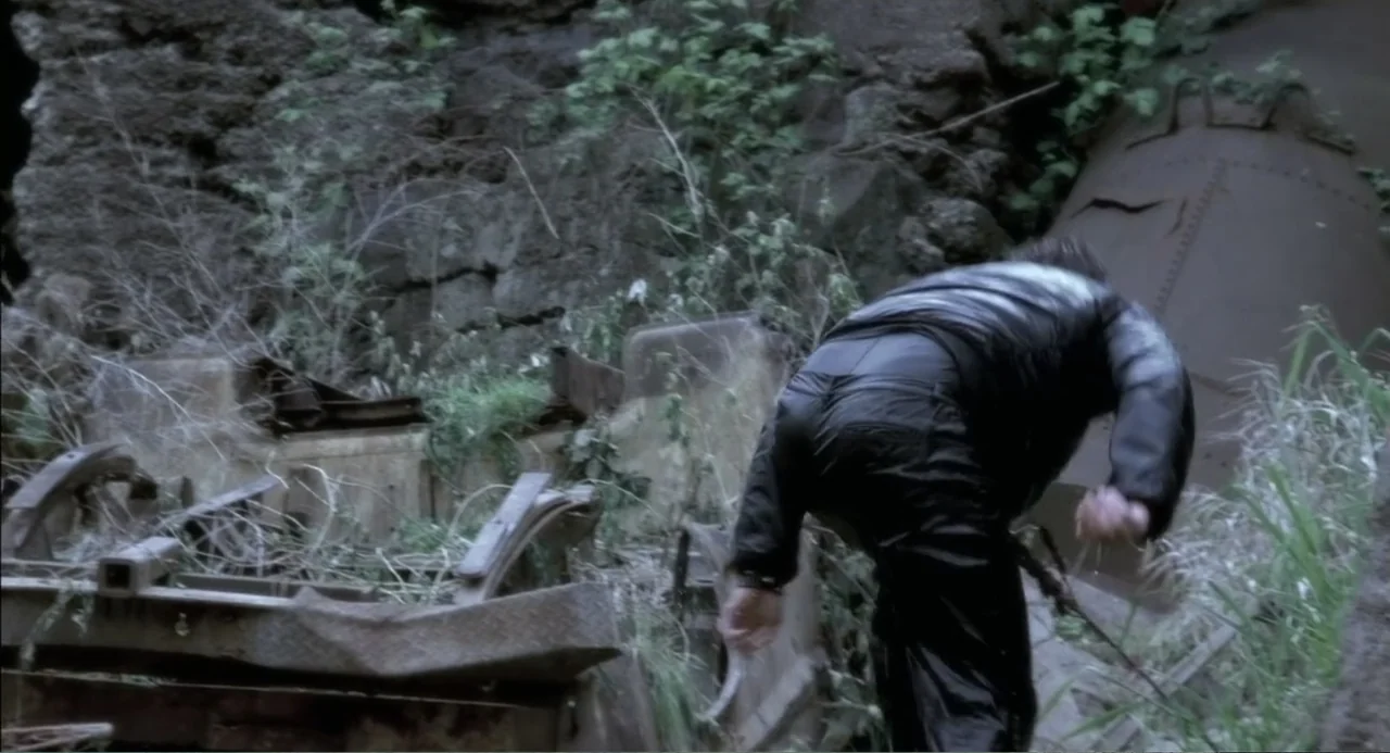 The Hunted (2003) movie still: medium — An overgrown, industrial junkyard features rusted metal components and tangled weeds among rock formations.…