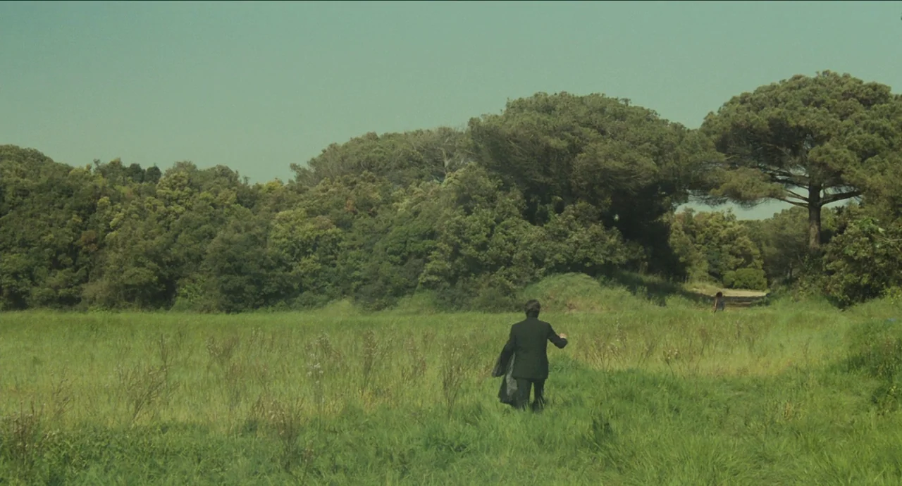 City of Women (1980) movie still: wide — A man walks across a sprawling green meadow towards a dense thicket of trees. The landscape is open and…