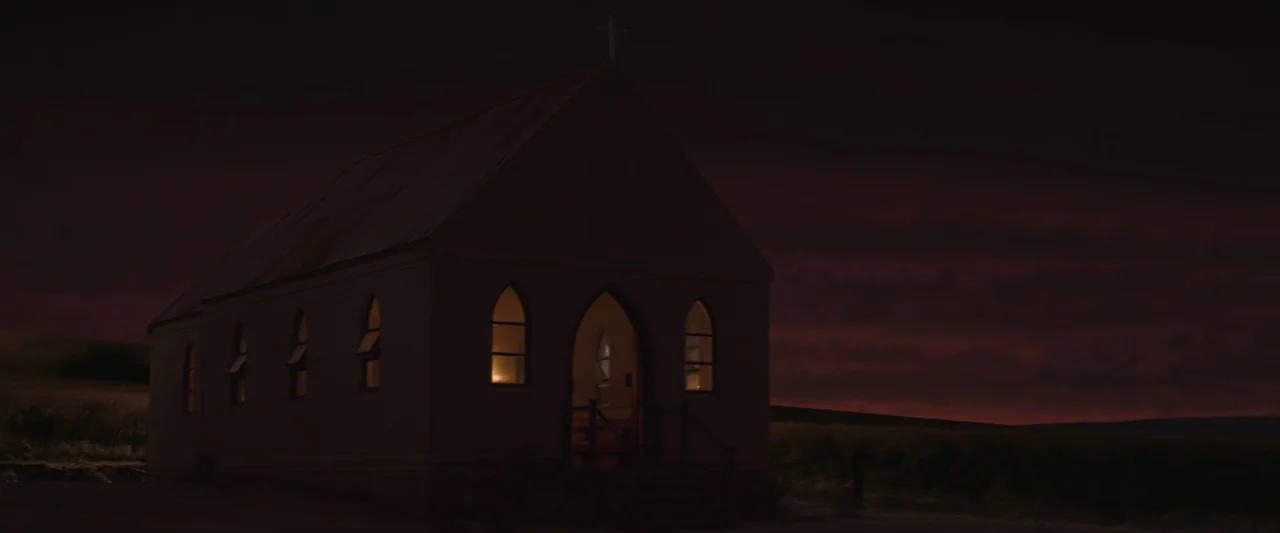 The New Boy (2023) movie still: wide — A small isolated church sits in a dark, empty field under a dusky sky. Warm light glows through the arched…
