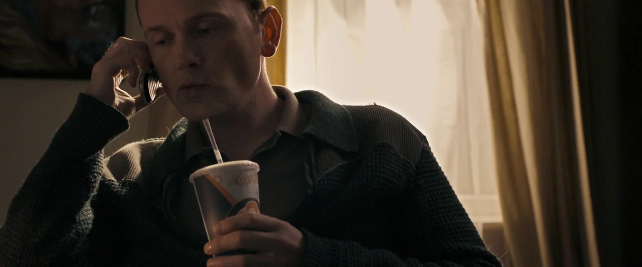 Compliance (2012) movie still: medium — An interior space features beige curtains illuminated by daylight from an adjacent window. A man in a dark…