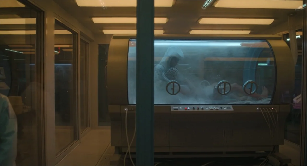 Mickey 17 (2025) movie still: medium — A sterile medical room features a large cylindrical pressurized tank filled with white vapor. A person in a…