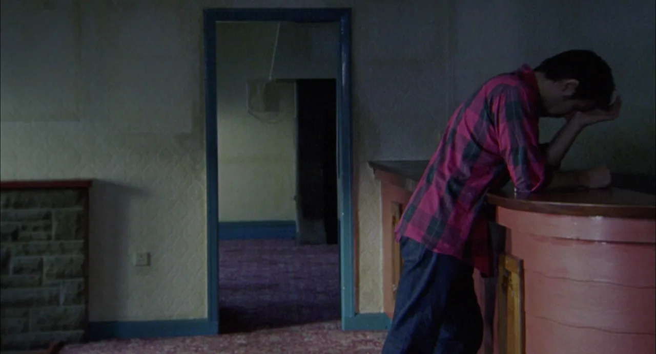 My Summer of Love movie still: medium — A dimly lit room with patterned walls and a doorway leading to darkness. A man in a flannel shirt leans…