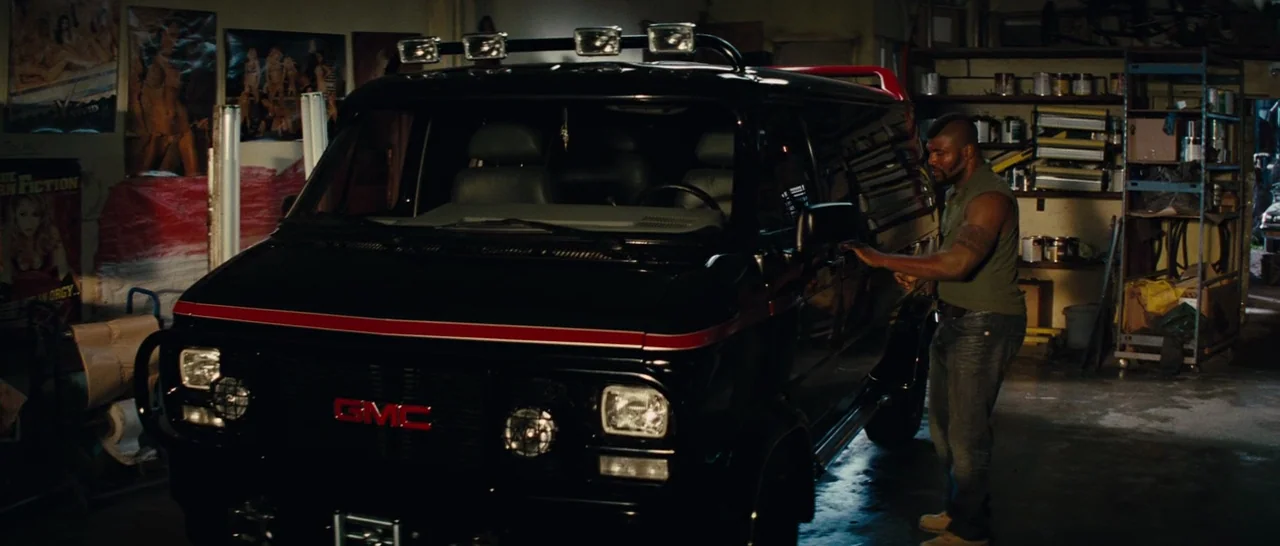 The A-Team (2010) movie still: medium — A cluttered workshop contains storage shelves, posters on the walls, and a large black van. A man wearing a…