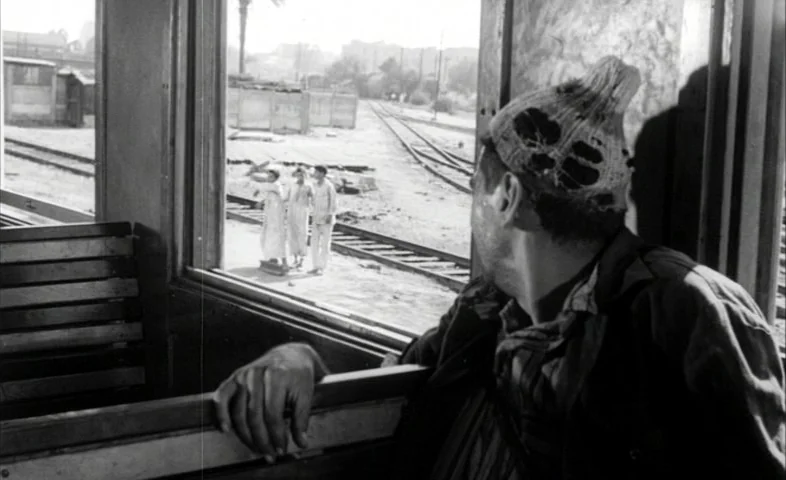 Cairo Station (1958) movie still: medium — A man sits inside a wooden passenger train carriage looking out an open window. Outside on the tracks…
