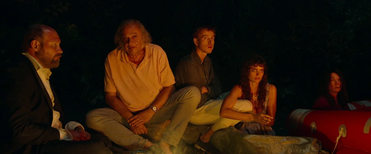 Triangle of Sadness (2022) movie still: medium — A group of five people sits in the dark on a beach around a fire. The orange light highlights their…