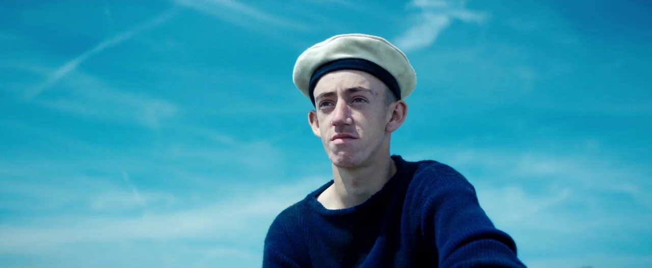 Slack Bay (2016) movie still: medium — A clear bright blue sky serves as the background for a portrait. A young man wearing a wool sweater and beret…