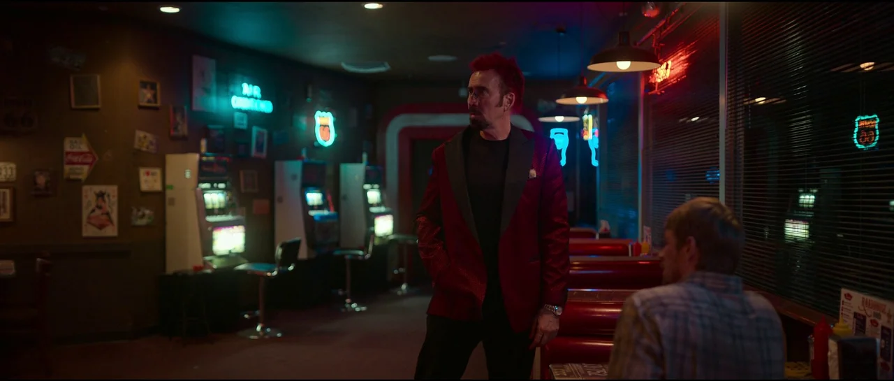 Sympathy For The Devil (2023) movie still: medium — A dimly lit diner interior with rows of booths and glowing neon wall signs. A man with red hair…