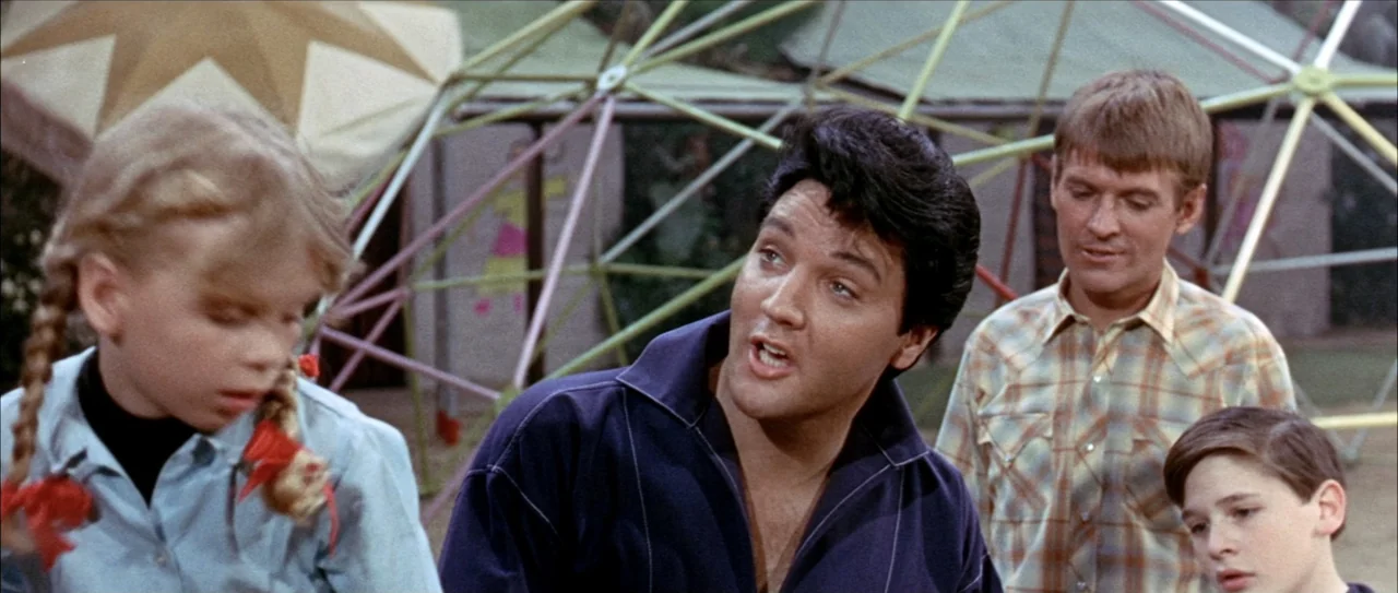 Clambake (1967) movie still: medium — A playground features a geometric jungle gym structure made of metal bars. A man talks to a young girl and a…
