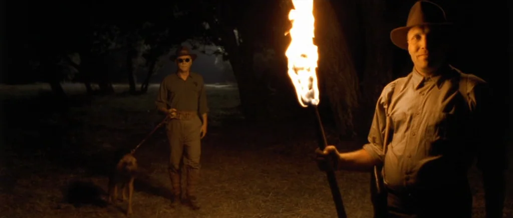 O Brother, Where Art Thou? (2000) movie still: medium — A dark clearing at night is surrounded by silhouetted trees and dense foliage. One man holds…