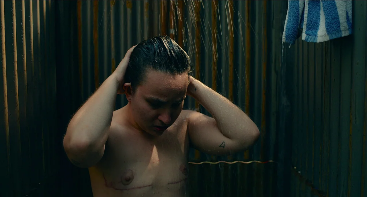 Trim Season (2023) movie still: medium — An outdoor shower area features a vertical corrugated metal wall with rust markings. A person stands…