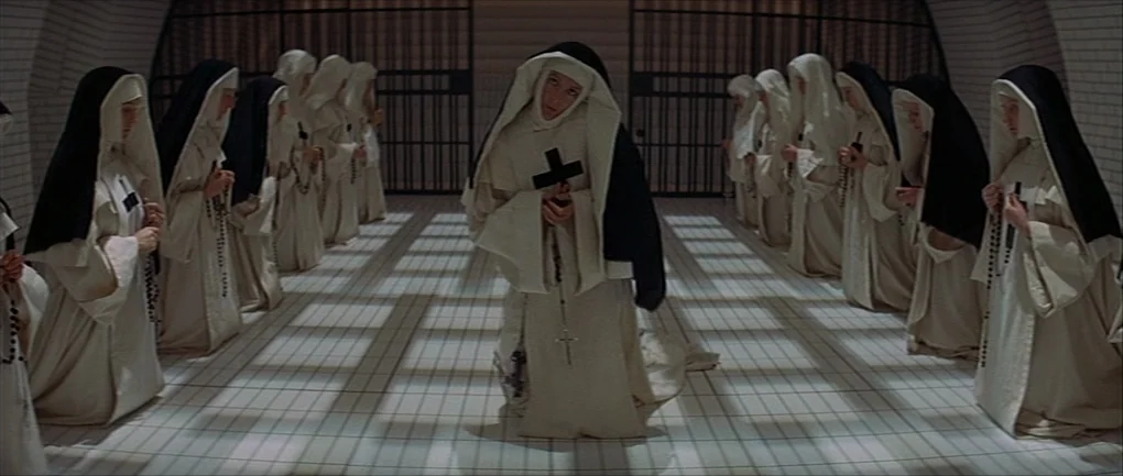 The Devils (1971) movie still: fullbody — A sterile room with white floor tiles and dark barred metal doors at ends. Multiple nuns wearing…