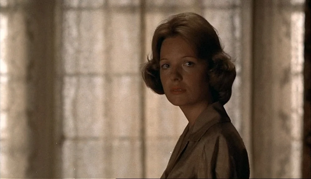 The Godfather (1972) movie still: closeup — A woman stands in front of a window covered by light linen curtains. The woman turns her head slightly…