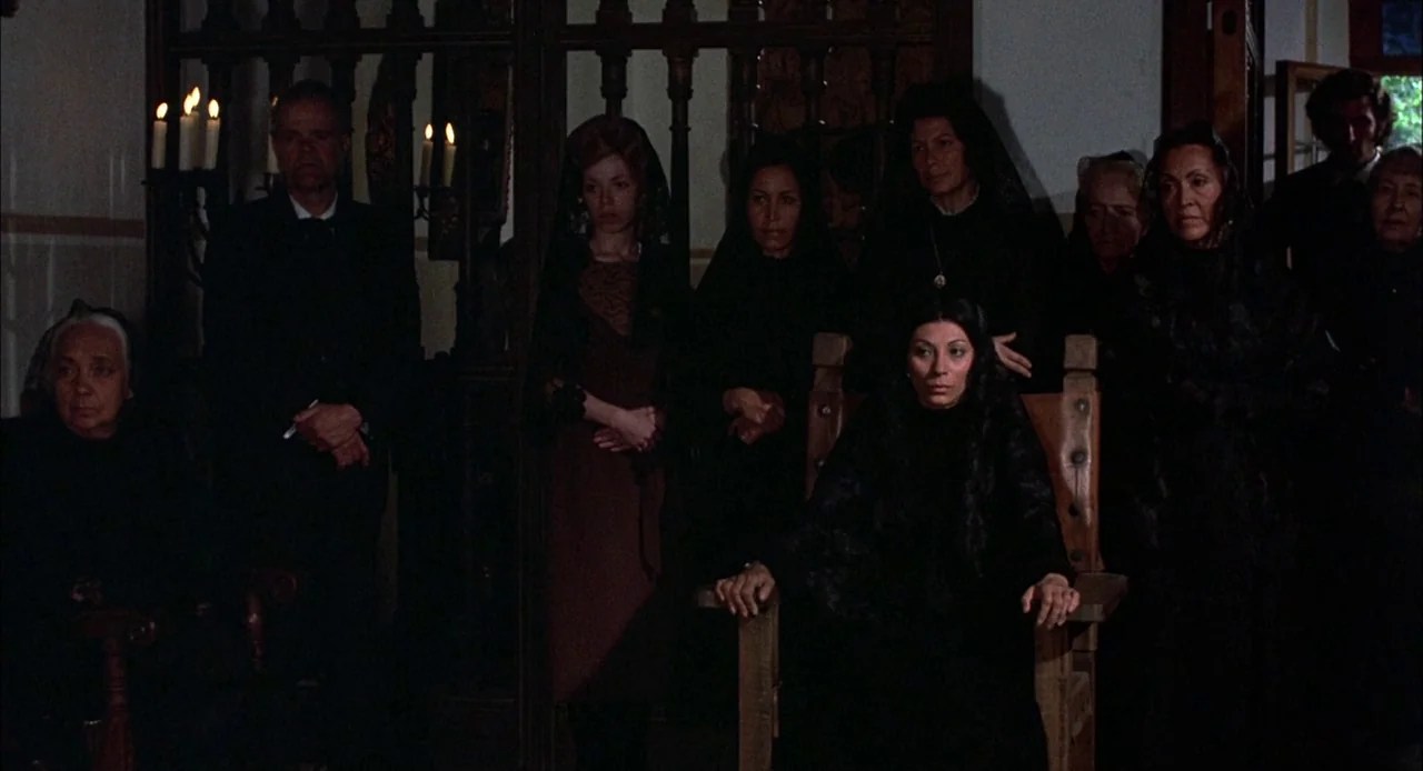 Bring Me The Head of Alfredo Garcia (1974) movie still: wide — A group of people wearing black mourning clothes gathers in a dim room. A woman sits…
