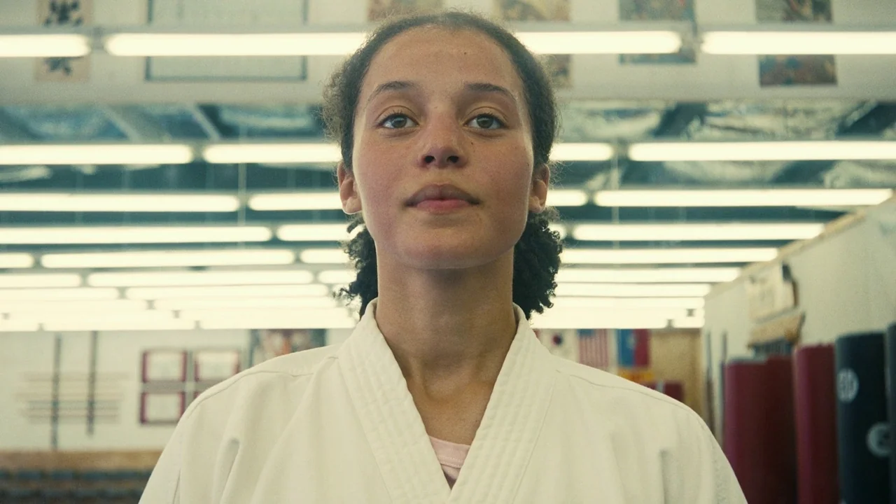 One Battle After Another (2025) movie still: medium — A well-lit martial arts training hall with rows of overhead lights. A young woman stands…