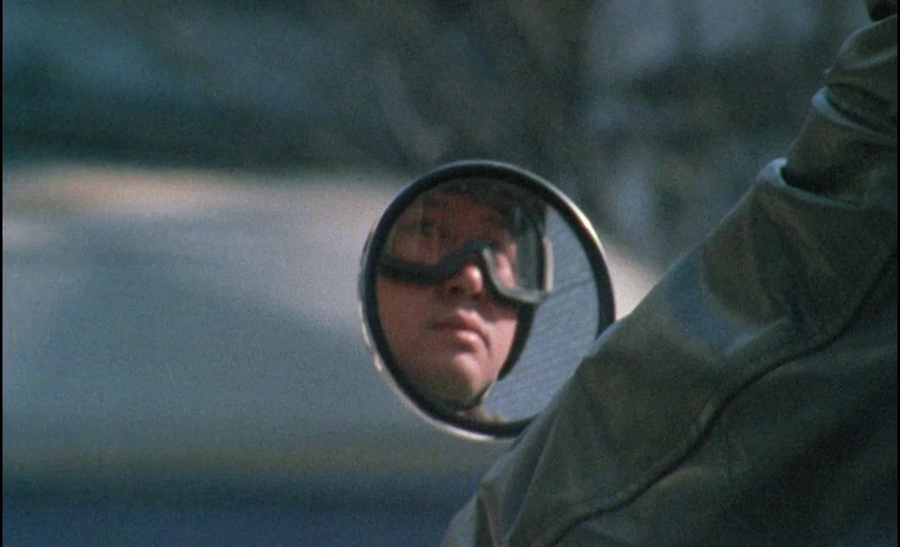 Sans Soleil (1983) movie still: closeup — A close-up view of a circular motorcycle mirror reflecting a male rider's face. The rider wears protective…
