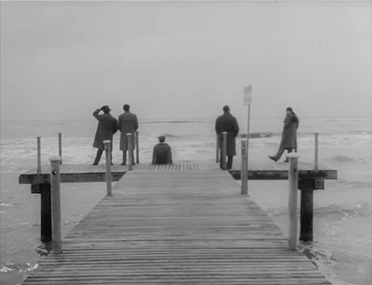 I Vitelloni (1953) movie still: wide — A wooden pier extends into a calm, grey sea under an overcast sky. Five men in long coats stand at the end of…