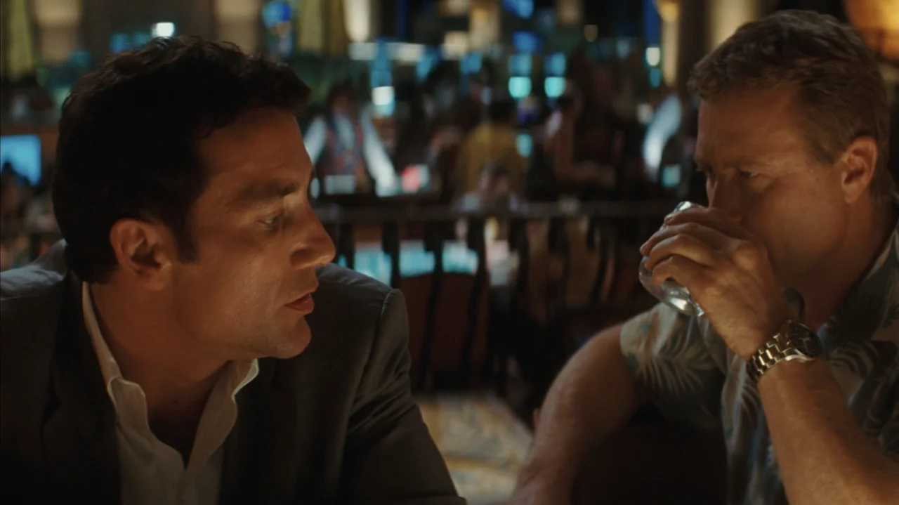 Duplicity (2009) movie still: medium — A dimly lit casino bar features a blurred background of slot machine displays. Two men sit at a counter, one…
