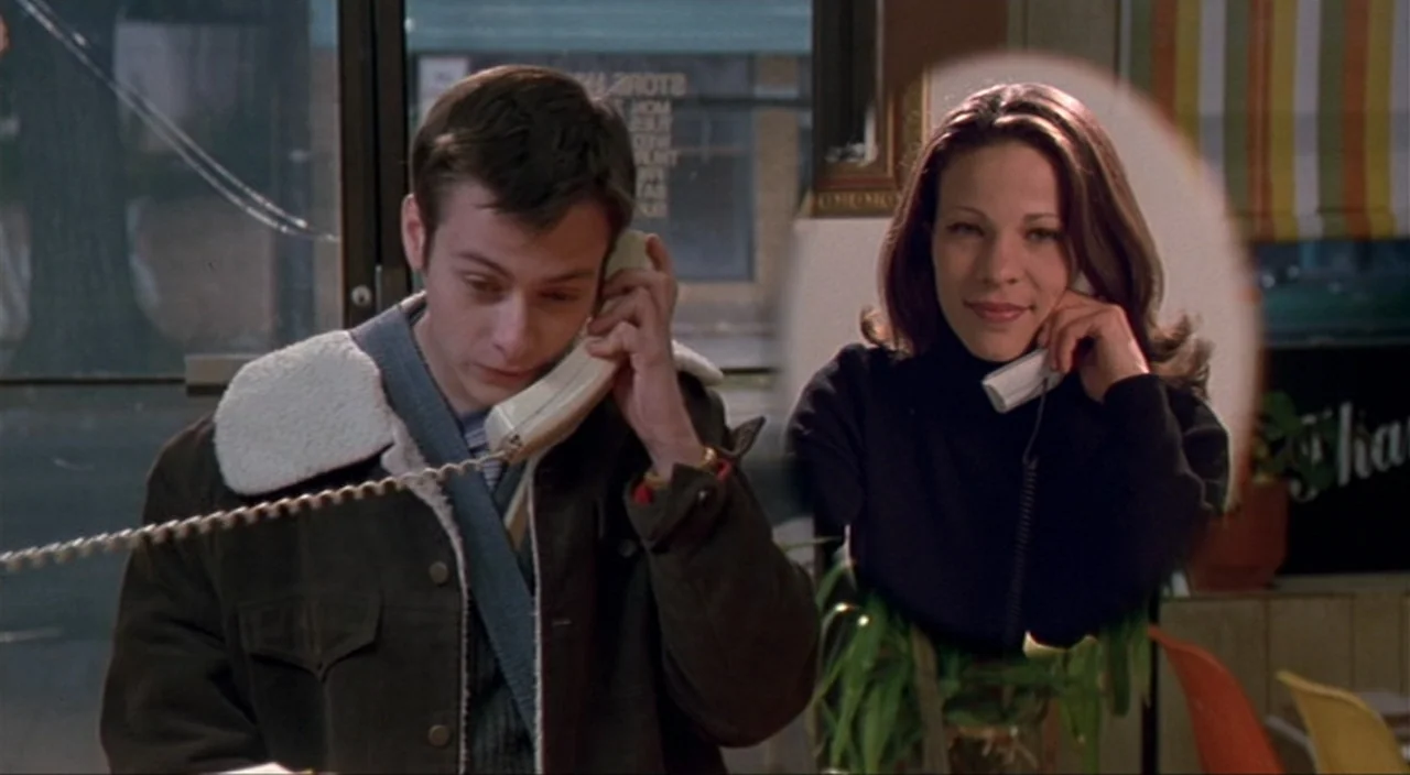 Pecker movie still: medium — A man speaks into a beige landline telephone inside a diner. A woman appears as a floating, semi-transparent image next…