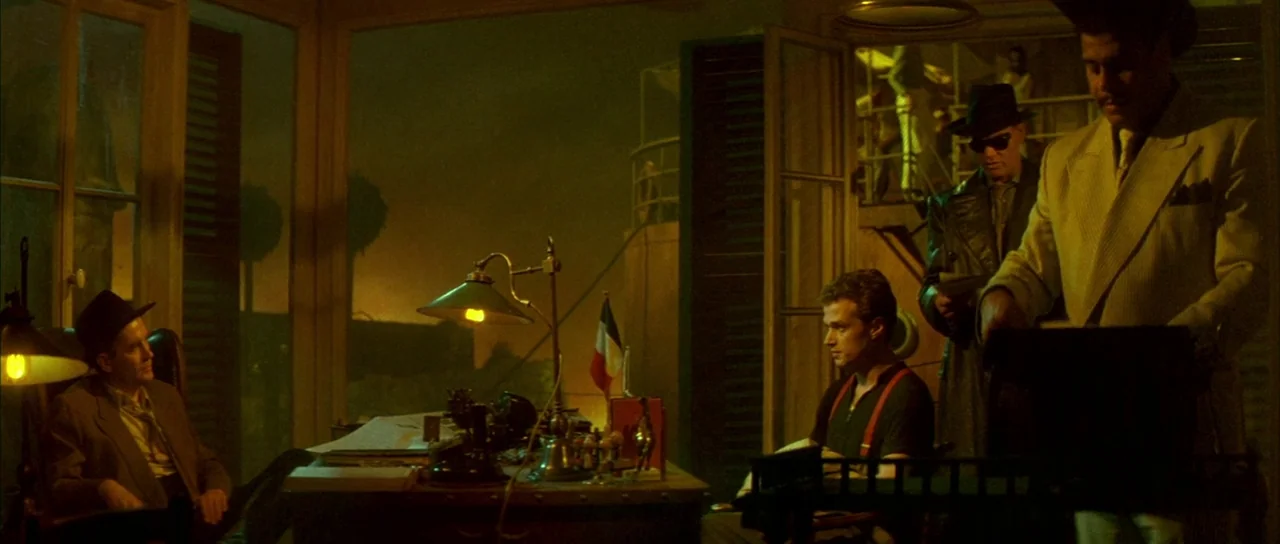 Querelle (1982) movie still: wide — An office at night with large windows overlooking a dark landscape with glowing distant lights. Four men stand…