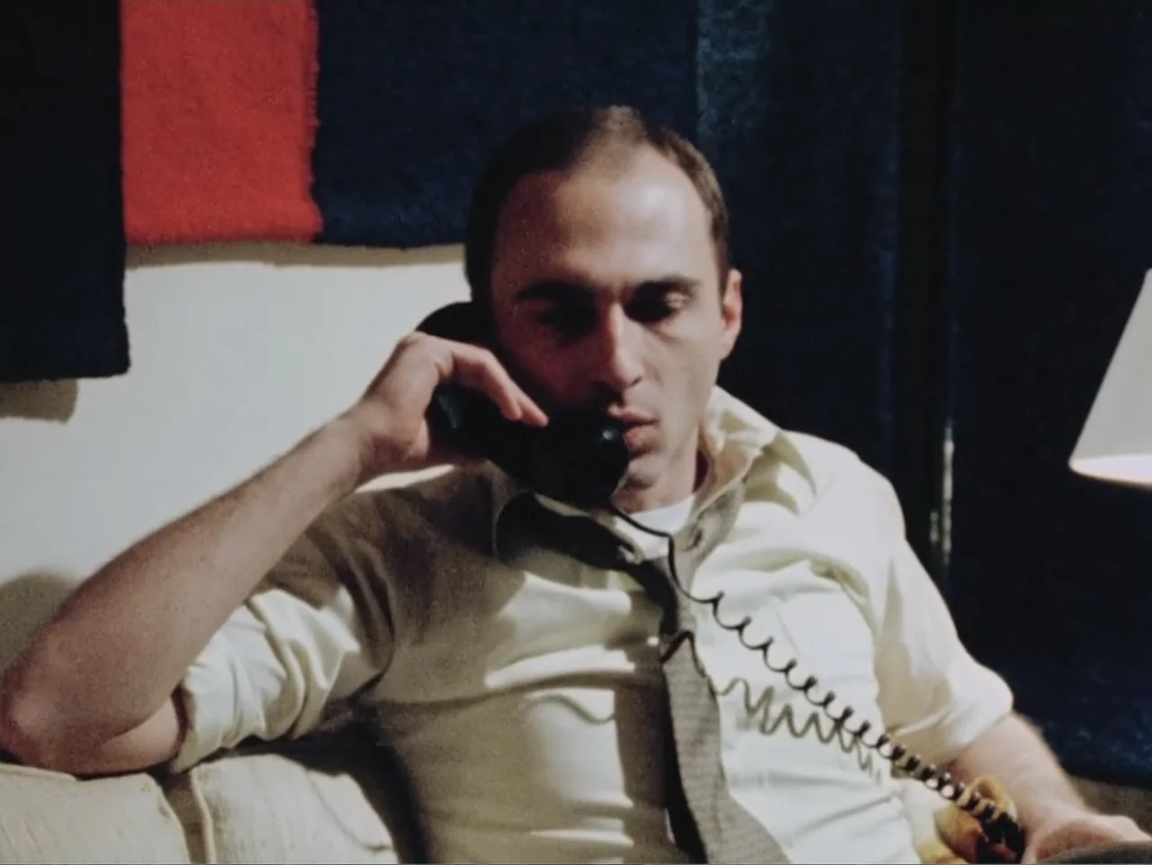 Born in Flames (1983) movie still: medium — A 1970s interior with a white wall decorated with dark and red panels. A man sits on a couch while…