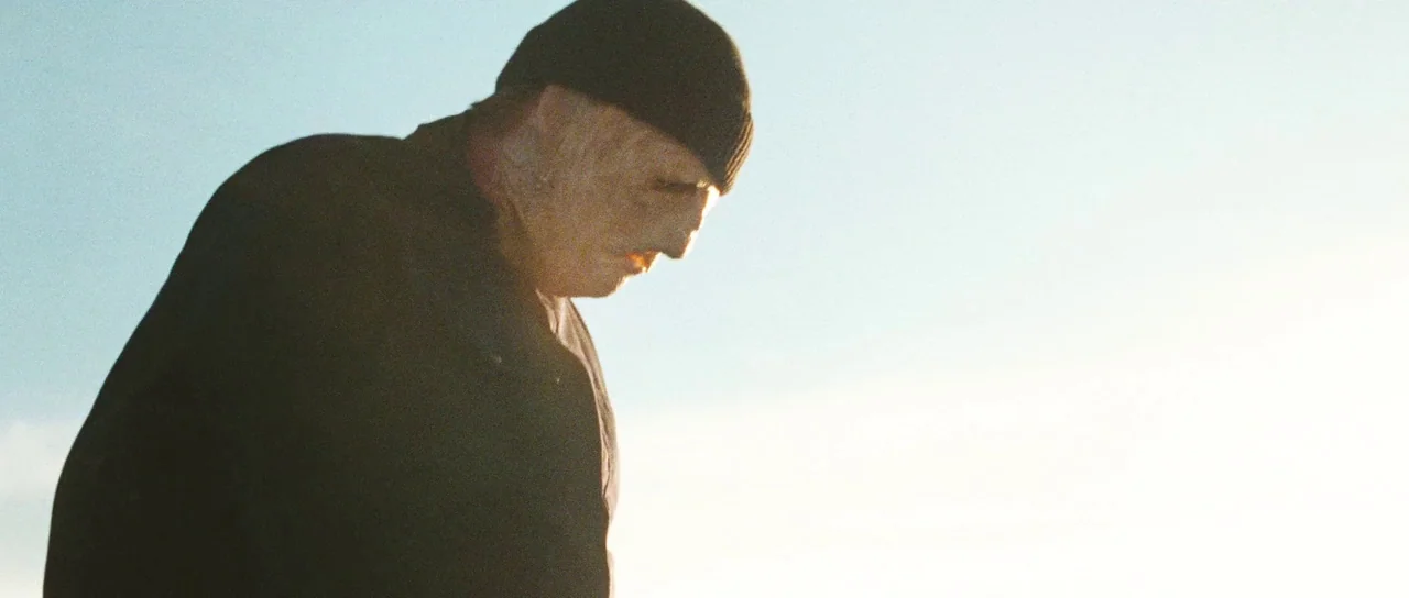 Surveillance (2008) movie still: medium — A man stands outdoors under an expansive, brightly lit white sky. The man wears a black hat and coat while…