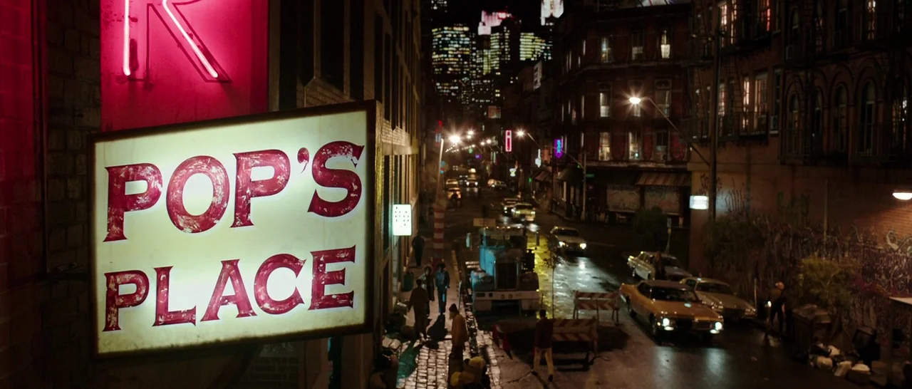 Predestination (2014) movie still: wide — A view of a nighttime urban street with a prominent illuminated sign hanging overhead. Cars are parked…