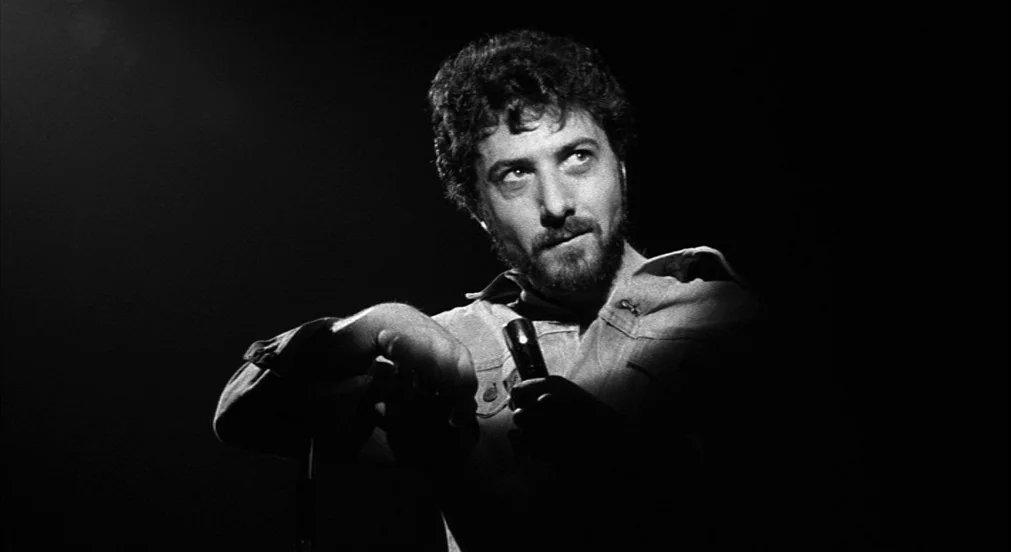 Lenny (1974) movie still: medium — A dark stage with no visible background elements beyond the spotlight-lit performer. A bearded man stands in the…