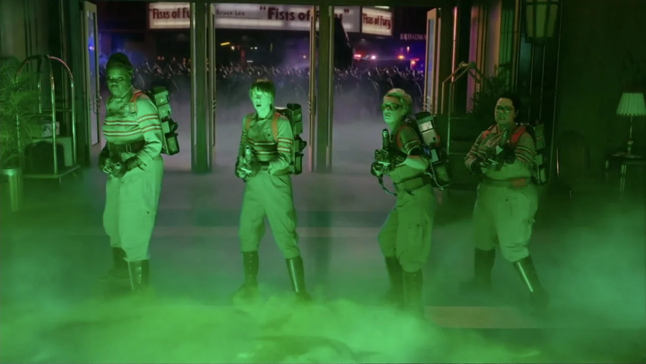 Ghostbusters (2016) movie still: fullbody — Four individuals wearing tan tactical jumpsuits stand together in a lobby filled with green smoke. They…
