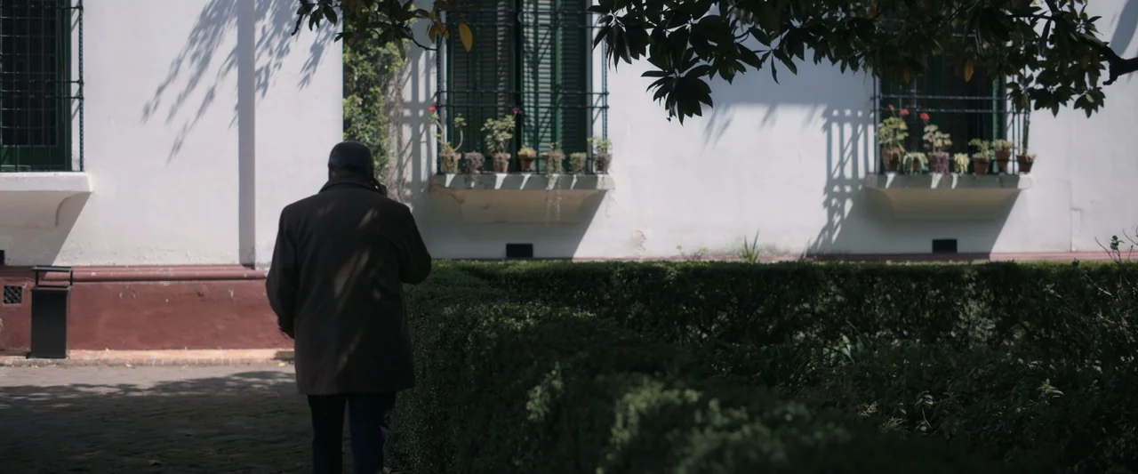 Doublespeak (2021) movie still: medium — A man walks past a manicured hedge in front of a white building facade. The building features shuttered…