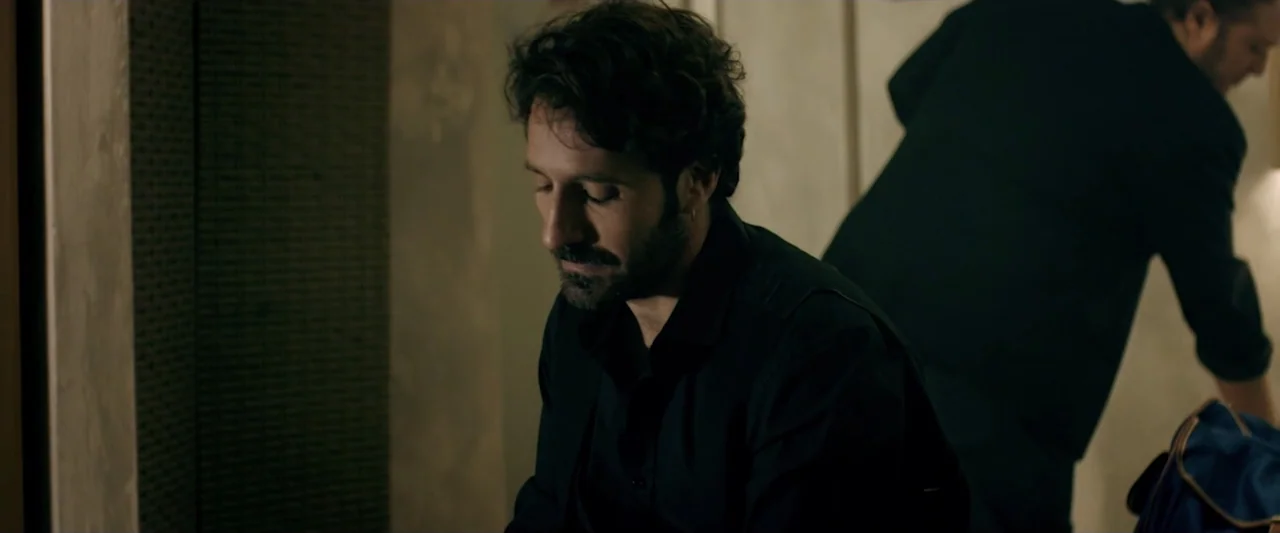 Taranta On the Road (2017) movie still: medium — A dimly lit room features a man sitting quietly in the foreground. A second figure stands partially…