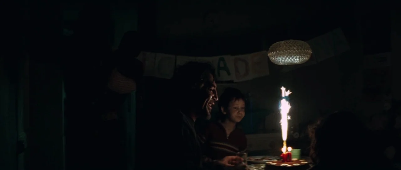 Biutiful (2010) movie still: medium — A dimly lit room contains a table with a birthday cake and sparkler. A man and a child sit at the table…