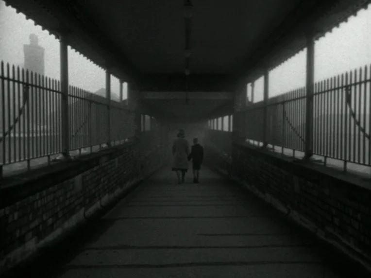 Death and Transfiguration (1983) movie still: wide — An enclosed metal and brick pedestrian bridge crosses above a misty urban area. A woman and a…