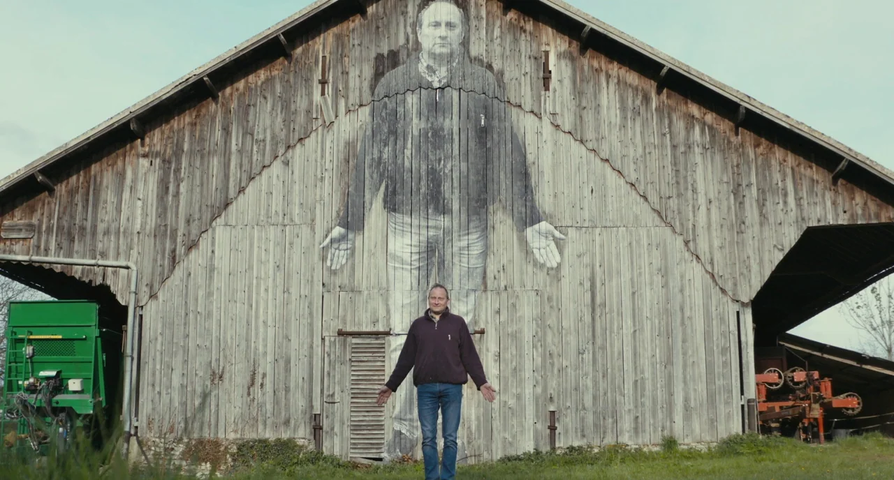 Faces Places (2017) movie still: fullbody — An aging wooden structure displays a massive mural of a man on its facade. A real man stands in front,…