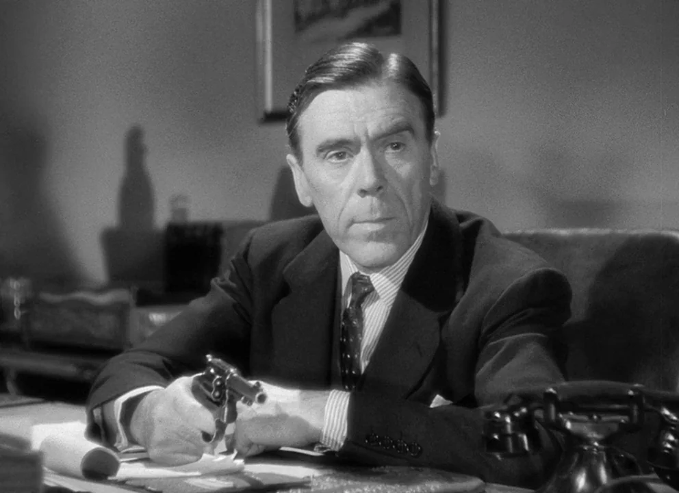 Spellbound (1945) movie still: medium — A dimly lit room with a man sitting behind a dark wooden desk. The man holds a small firearm pointed…