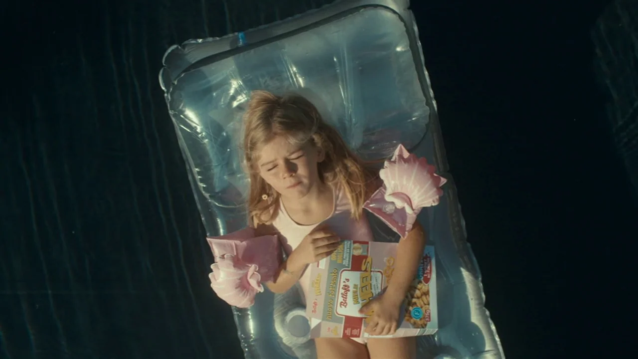 Amanda (2022) movie still: medium — A young girl floats on a clear plastic raft in deep blue water. She holds a box of cereal to her chest while…