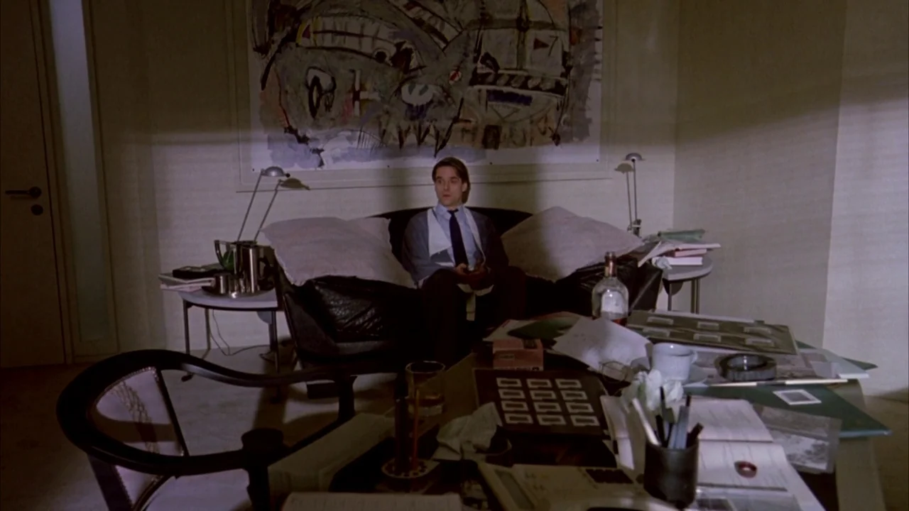 Dead Ringers (1988) movie still: wide — A cluttered room features a large abstract painting hanging above a dark leather sofa. A man sits on the…