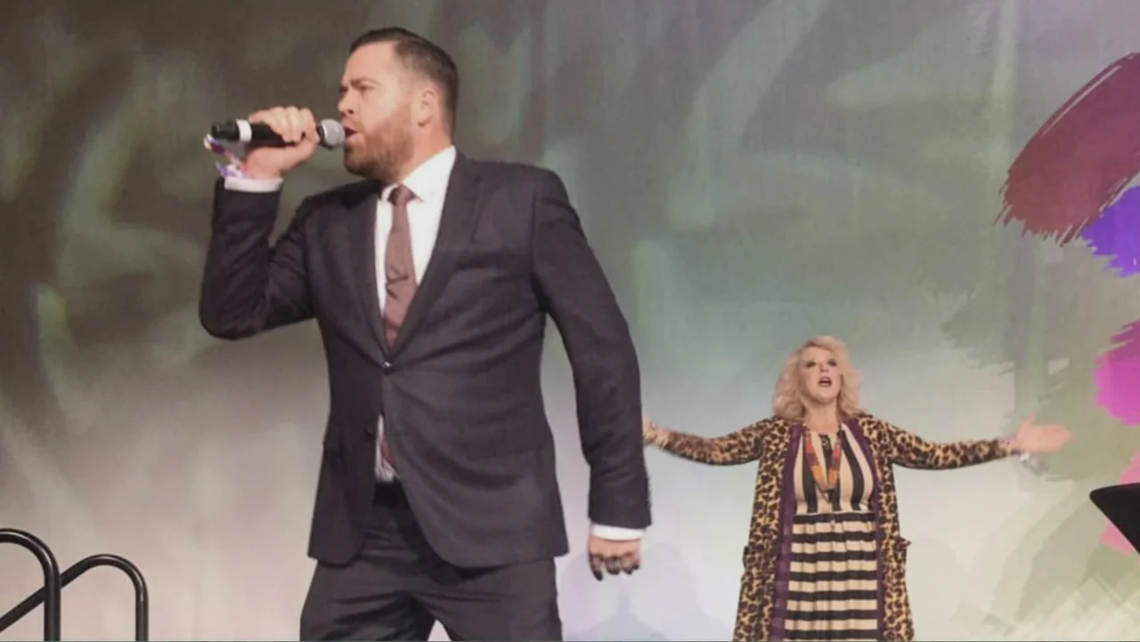 The Rise and Fall of Lularoe (2021) movie still: medium — A man and a woman stand on a brightly lit indoor stage area. The man performs with a…