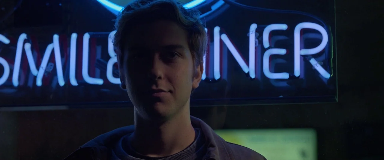 Death Note (2017) movie still: medium — A young man stands behind a glass window at night. A bright blue neon sign glows overhead.