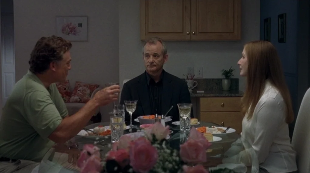 Broken Flowers movie still: medium — A residential dining room features a round glass table with plates and glasses. Three adults sit at the table…