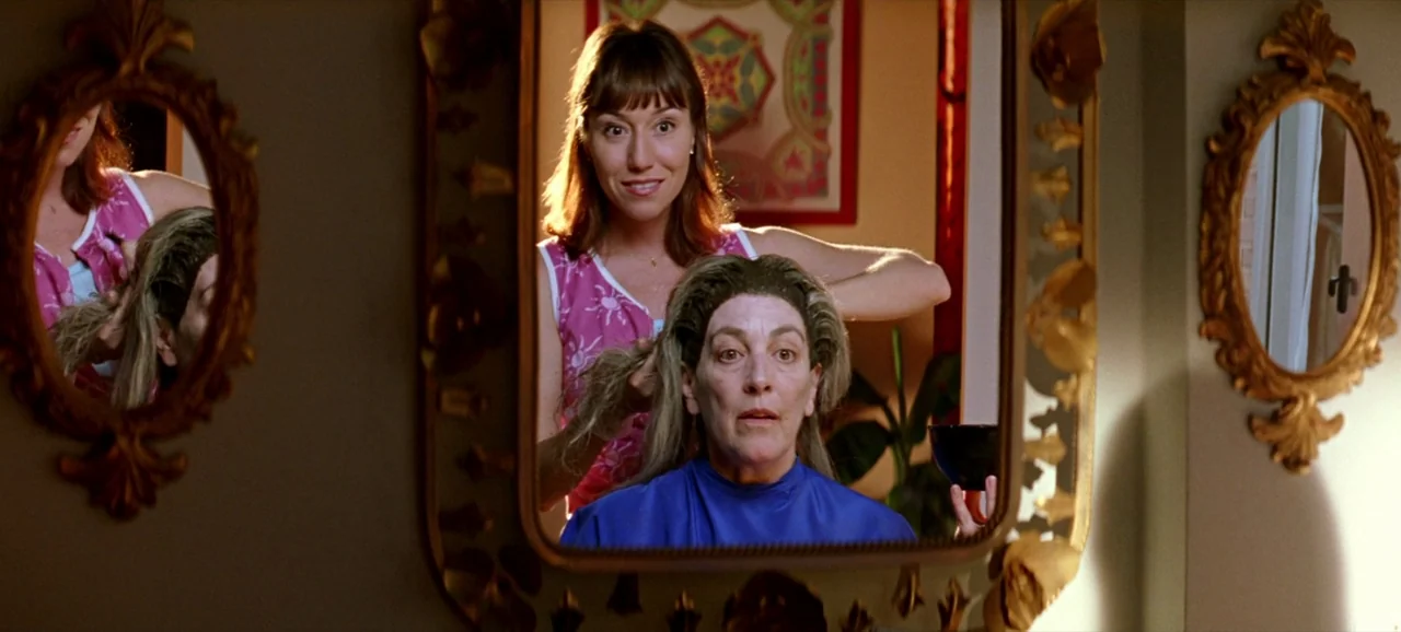 Volver (2006) movie still: medium — A woman stands behind another seated woman who is wearing a salon cape. The reflection of both women is visible…
