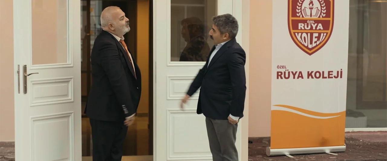Wrong Package (2025) movie still: fullbody — A modern building exterior features a white double door and an orange educational banner. Two men in…