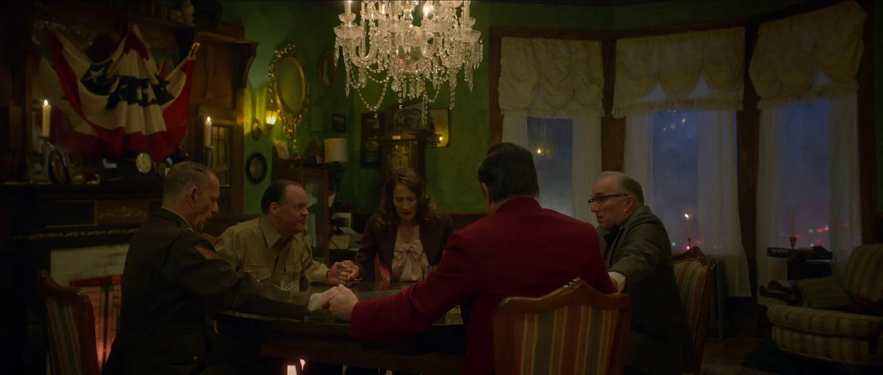 Brooklyn 45 (2023) movie still: wide — A green-walled dining room features a hanging crystal chandelier above a wooden table. Five people in period…