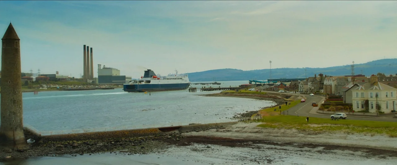Unwelcome (2022) movie still: establishing — A coastal landscape features a tall stone tower, a large ferry, and town buildings. The scene shows a…