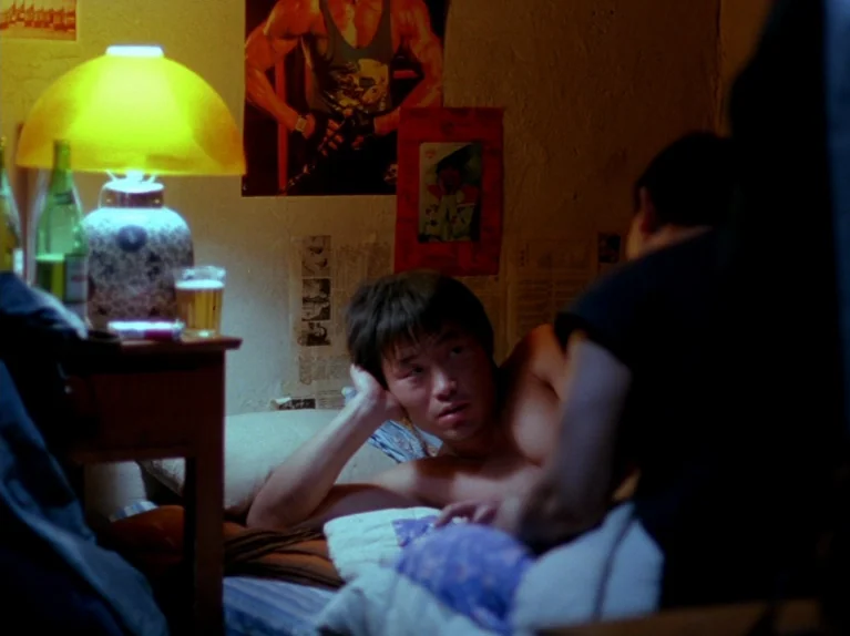 Black Snow (1990) movie still: medium — A small room features posters on the wall and a desk with a lamp. A man reclines on a bed while facing…